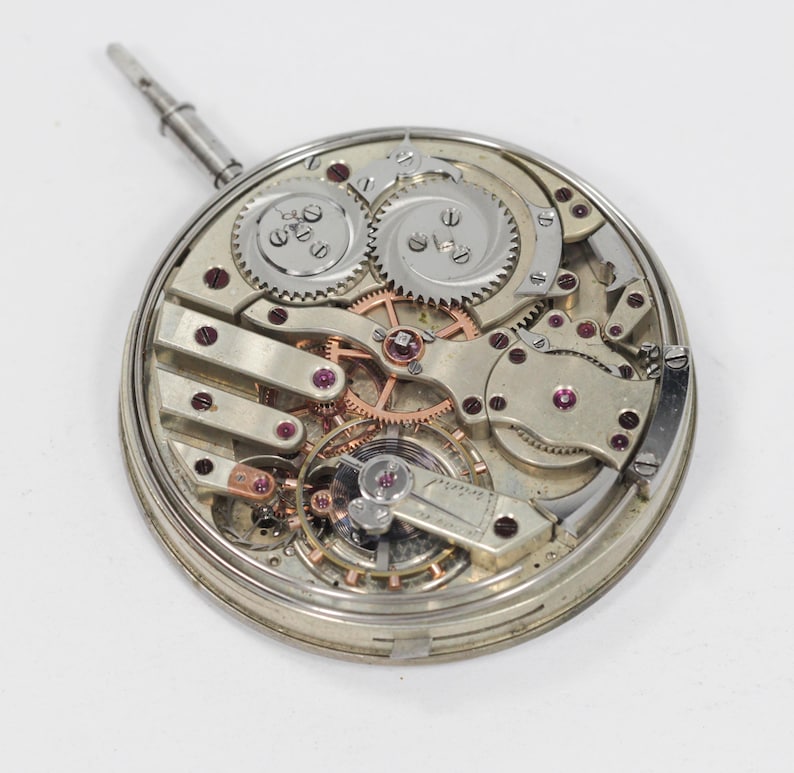 Puede incluir: Un movimiento de reloj plateado con engranajes, resortes y joyas de rub&iacute; expuestos. El intrincado mecanismo presenta m&uacute;ltiples capas de componentes, incluido un v&aacute;stago de cuerda. El reloj est&aacute; sobre un fondo blanco.