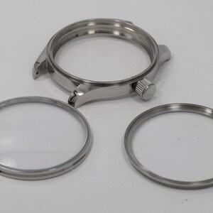 May include: A disassembled watch with a silver-colored case, a bezel, and two silver rings. The watch case has a crown and lugs for attaching a strap. The components are arranged on a white surface.