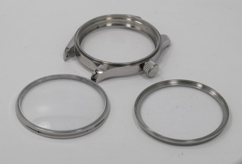 May include: A disassembled watch with a silver-colored case, a bezel, and two silver rings. The watch case has a crown and lugs for attaching a strap. The components are arranged on a white surface.