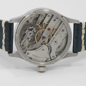 May include: Close-up of a silver-toned wristwatch with a visible mechanical movement. The watch has a dark blue leather strap with white stitching. The watch face displays intricate gears and the text "INTERNATIONAL WATCH CO."