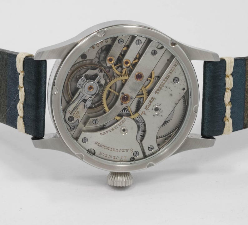 May include: Close-up of a silver-toned wristwatch with a visible mechanical movement. The watch has a dark blue leather strap with white stitching. The watch face displays intricate gears and the text "INTERNATIONAL WATCH CO."