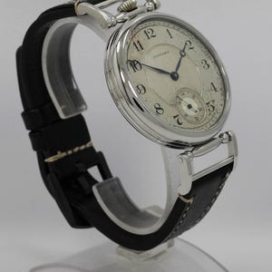 K&ouml;nnte beinhalten: Eine silberfarbene Armbanduhr mit rundem, wei&szlig;em Zifferblatt, schwarzen arabischen Ziffern, blauen Zeigern und dem Schriftzug Howard. Die Uhr verf&uuml;gt &uuml;ber ein strukturiertes Zifferblattzentrum, ein kleines Sekunden-Hilfszifferblatt und ein schwarzes Lederarmband.