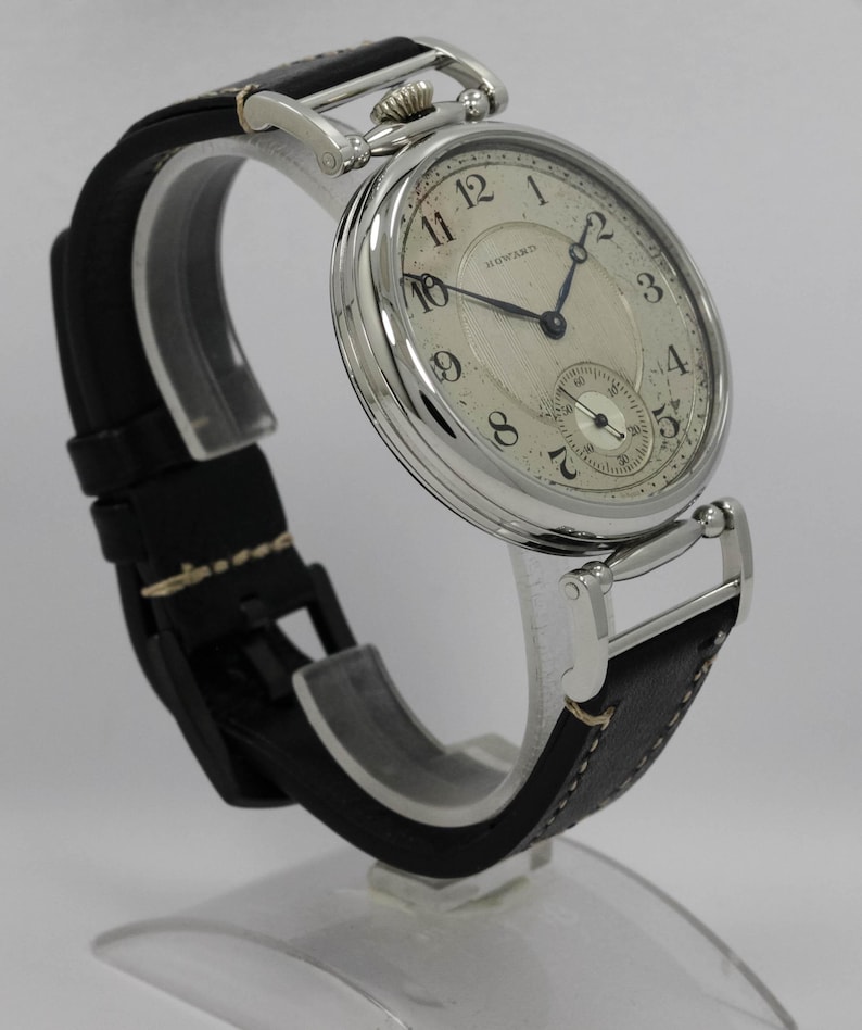 K&ouml;nnte beinhalten: Eine silberfarbene Armbanduhr mit rundem, wei&szlig;em Zifferblatt, schwarzen arabischen Ziffern, blauen Zeigern und dem Schriftzug Howard. Die Uhr verf&uuml;gt &uuml;ber ein strukturiertes Zifferblattzentrum, ein kleines Sekunden-Hilfszifferblatt und ein schwarzes Lederarmband.