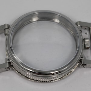 May include: A stainless steel watch case with a clear crystal. The case has a round shape with a ridged bezel and integrated lugs for the watch band. The watch case is designed to hold the watch movement and protect the watch face.