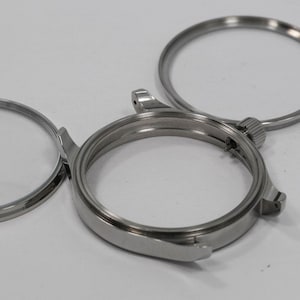 May include: Close-up of disassembled watch components. The image shows a silver-colored watch case, a bezel, and a crystal. The parts are arranged on a white surface, highlighting the intricate details of the watchmaking process.