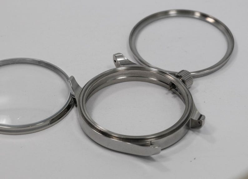 May include: Close-up of disassembled watch components. The image shows a silver-colored watch case, a bezel, and a crystal. The parts are arranged on a white surface, highlighting the intricate details of the watchmaking process.