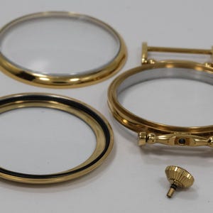 May include: Gold-toned watch parts, including a bezel, a crystal, and a stem. The parts are laid out on a white surface.