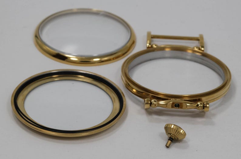 May include: Gold-toned watch parts, including a bezel, a crystal, and a stem. The parts are laid out on a white surface.