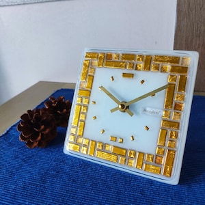 May include: White square clock with gold mosaic design and black hands. The clock face has the text "Ponyglass" on the bottom right corner.