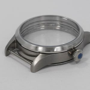 May include: A silver-toned watch case with a clear crystal and a blue-accented crown. The watch case has a brushed metal finish and a circular design, showcasing the watch's internal components.