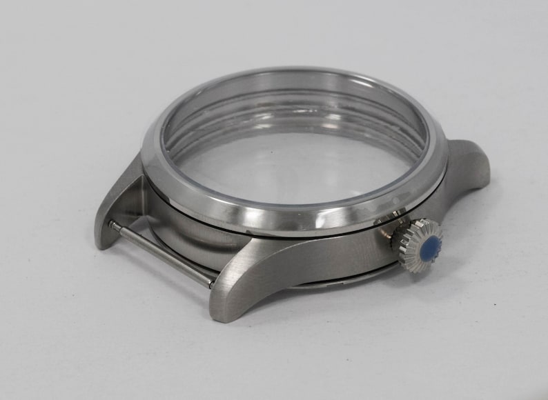 May include: A silver-toned watch case with a clear crystal and a blue-accented crown. The watch case has a brushed metal finish and a circular design, showcasing the watch's internal components.