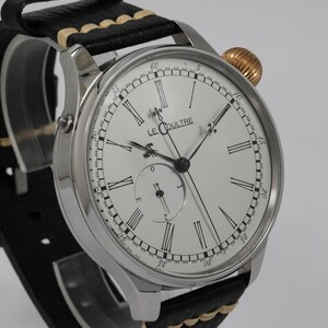 May include: A silver wristwatch with a white face and black Roman numeral hour markers. The watch has a black leather strap with a tan stitching detail. The watch face also features the text "LE COULTRE".