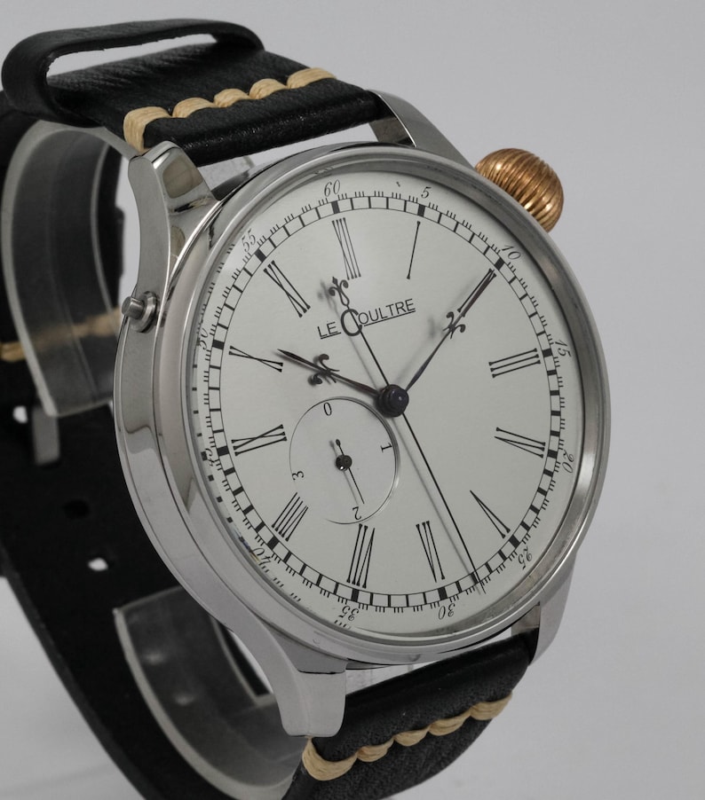 May include: A silver wristwatch with a white face and black Roman numeral hour markers. The watch has a black leather strap with a tan stitching detail. The watch face also features the text "LE COULTRE".