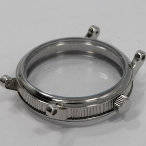 May include: A silver-toned watch case with a clear glass face. The case has a ridged band and small lugs for attaching a watch strap. A small, round knob is visible on the side.