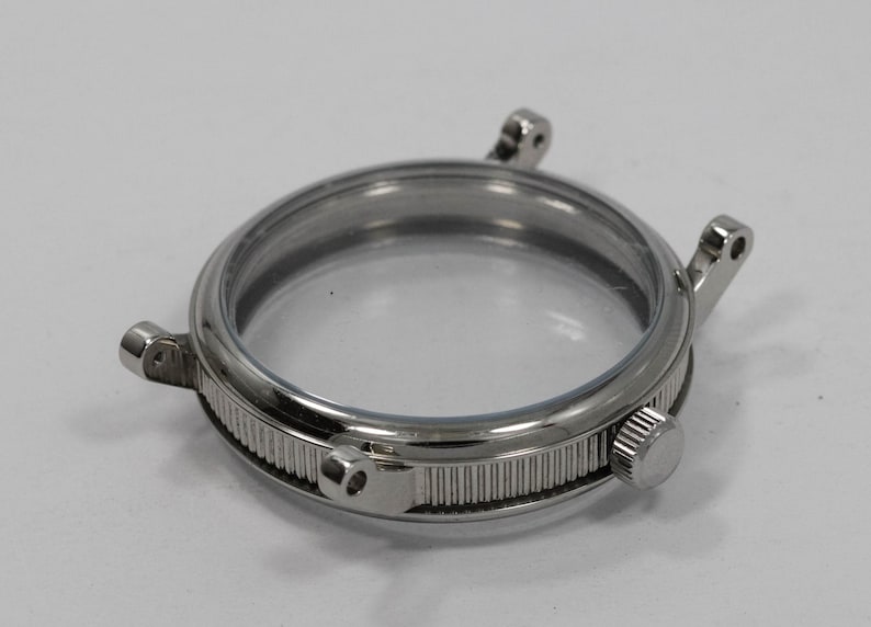 May include: A silver-toned watch case with a clear glass face. The case has a ridged band and small lugs for attaching a watch strap. A small, round knob is visible on the side.