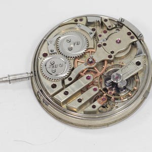 Puede incluir: Primer plano del mecanismo de un reloj antiguo, que muestra intrincados engranajes, muelles y joyas. Los componentes plateados est&aacute;n dispuestos en un dise&ntilde;o circular, con un v&aacute;stago que se extiende desde el lateral. El mecanismo del reloj es un testimonio de la ingenier&iacute;a de precisi&oacute;n.