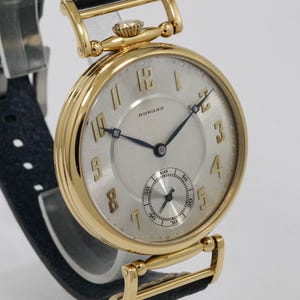 May include: A gold wristwatch with a light-coloured face and black hands. The watch features a small seconds dial and the name "HOWARD" is printed on the face. The watch has a black leather strap.