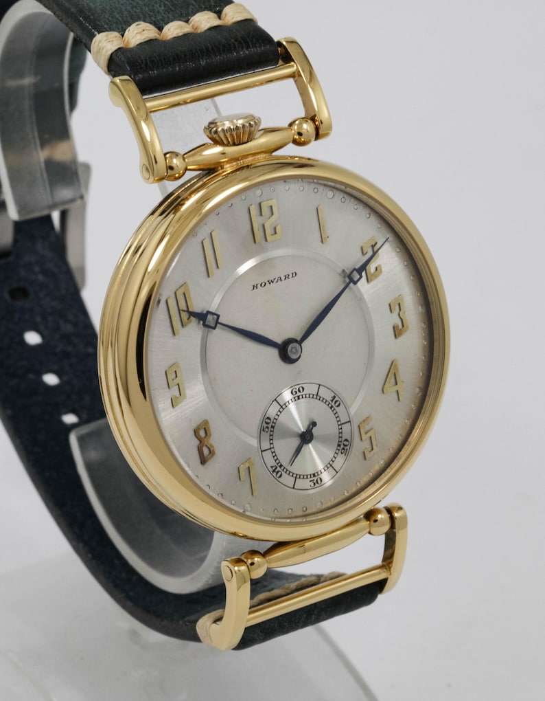 May include: A gold wristwatch with a light-coloured face and black hands. The watch features a small seconds dial and the name "HOWARD" is printed on the face. The watch has a black leather strap.