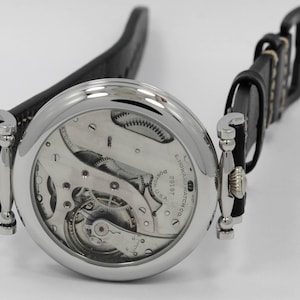 K&ouml;nnte beinhalten: Eine silberfarbene Armbanduhr mit schwarzem Lederarmband und skelettiertem Zifferblatt, das das mechanische Uhrwerk freilegt. Auf dem Zifferblatt sind der Text E. HOWARD WATCH CO. BOSTON U.S.A. und die Seriennummer 29197 eingraviert.