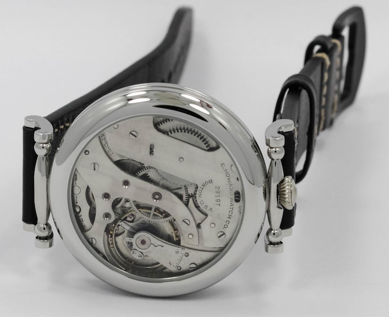 K&ouml;nnte beinhalten: Eine silberfarbene Armbanduhr mit schwarzem Lederarmband und skelettiertem Zifferblatt, das das mechanische Uhrwerk freilegt. Auf dem Zifferblatt sind der Text E. HOWARD WATCH CO. BOSTON U.S.A. und die Seriennummer 29197 eingraviert.