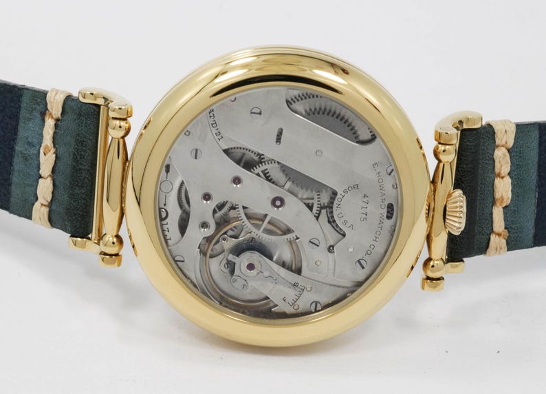 May include: A gold wristwatch showcasing its mechanical workings, with a dark green and blue striped leather strap. The watch face reveals intricate gears and the inscription "E. Howard Watch Co., Boston, U.S."