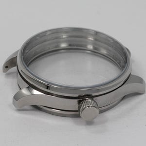 May include: A stainless steel watch case with a clear crystal and a crown. The watch case has a round shape with lugs for attaching a watch band. The case is designed to house the watch movement and protect the watch face.