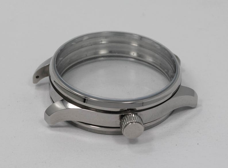 May include: A stainless steel watch case with a clear crystal and a crown. The watch case has a round shape with lugs for attaching a watch band. The case is designed to house the watch movement and protect the watch face.