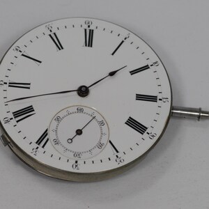 Puede incluir: Una esfera de reloj blanca con n&uacute;meros romanos y manecillas negras. Una esfera m&aacute;s peque&ntilde;a en la parte inferior. La esfera tiene una caja de metal plateado y un v&aacute;stago plateado. La esfera es un componente de un reloj de bolsillo.