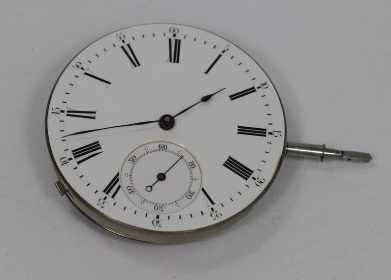 Puede incluir: Una esfera de reloj blanca con n&uacute;meros romanos y manecillas negras. Una esfera m&aacute;s peque&ntilde;a en la parte inferior. La esfera tiene una caja de metal plateado y un v&aacute;stago plateado. La esfera es un componente de un reloj de bolsillo.