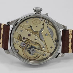 Puede incluir: Un reloj plateado con una correa de cuero marr&oacute;n. La parte posterior del reloj est&aacute; abierta, revelando el intrincado funcionamiento interno del movimiento. La esfera del reloj tiene el texto "VACHERON CONSTANTIN GENEVE" impreso en ella.