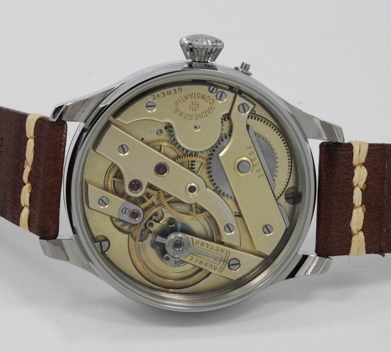 Puede incluir: Un reloj plateado con una correa de cuero marr&oacute;n. La parte posterior del reloj est&aacute; abierta, revelando el intrincado funcionamiento interno del movimiento. La esfera del reloj tiene el texto "VACHERON CONSTANTIN GENEVE" impreso en ella.