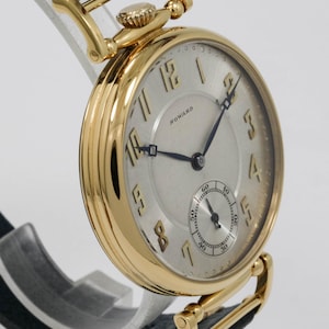 May include: A gold wristwatch with a silver face and black hands. The watch features a small seconds dial and the name "HOWARD" is printed on the face. It has a black leather strap with gold accents.