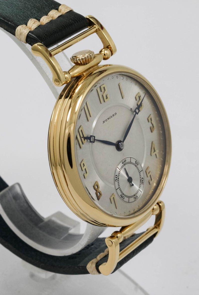May include: A gold wristwatch with a silver face and black hands. The watch features a small seconds dial and the name "HOWARD" is printed on the face. It has a black leather strap with gold accents.