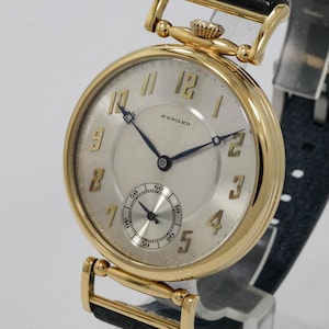 May include: A gold wristwatch with a cream face and black leather strap. The watch face displays gold numerals, a small seconds dial, and the brand name "HOWARD". The watch has a classic, vintage design.