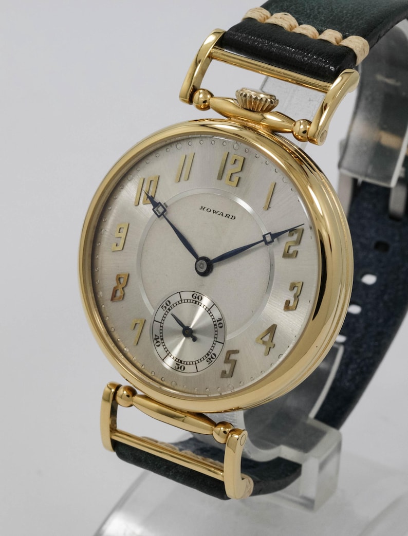 May include: A gold wristwatch with a cream face and black leather strap. The watch face displays gold numerals, a small seconds dial, and the brand name "HOWARD". The watch has a classic, vintage design.