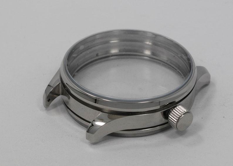 May include: A stainless steel watch case with a clear crystal and a silver crown. The watch case has a round shape with lugs for attaching a watch band. The case is designed to house the watch movement and protect the watch face.