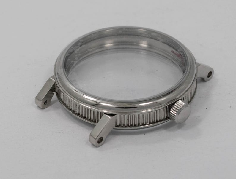 May include: A silver-toned watch case with a clear crystal. The case has a ridged bezel and two lugs for the watch band. A small, round crown is visible on the side. The watch case is designed to hold a watch movement.