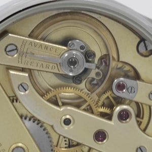 Puede incluir: Primer plano de un movimiento de reloj con engranajes de color dorado y una palanca plateada con la etiqueta "AVANCE" y "RETARD".
