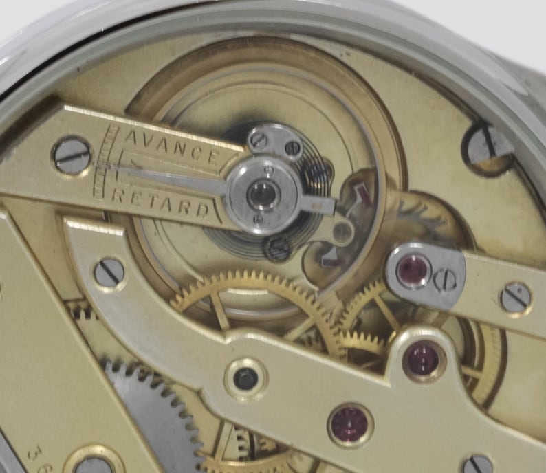 Puede incluir: Primer plano de un movimiento de reloj con engranajes de color dorado y una palanca plateada con la etiqueta "AVANCE" y "RETARD".