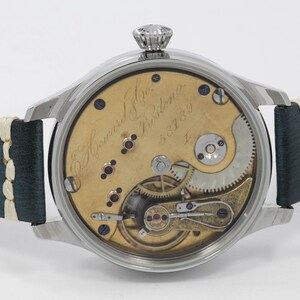May include: A detailed view of a vintage wristwatch, showcasing its internal mechanisms. The watch has a silver case and a leather strap. The gold-toned face displays intricate gears and springs. The watch is marked "E. Howard & Co. Boston".