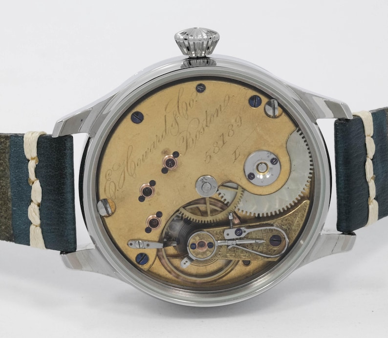 May include: A detailed view of a vintage wristwatch, showcasing its internal mechanisms. The watch has a silver case and a leather strap. The gold-toned face displays intricate gears and springs. The watch is marked "E. Howard & Co. Boston".