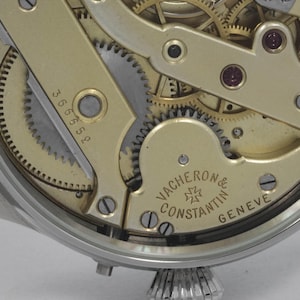 Puede incluir: Primer plano de un movimiento de reloj de tono dorado con engranajes intrincados y la inscripci&oacute;n "VACHERON CONSTANTIN GENEVE" en la parte inferior.