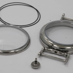 May include: A set of watch parts including a clear glass crystal, a metal case back, a metal strap, and a small metal screw.