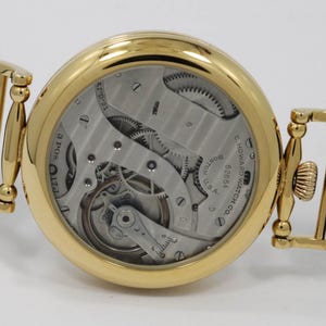 May include: A gold pocket watch with a black leather strap and a white stitching detail. The watch face is open, revealing the intricate inner workings of the watch. The watch is engraved with the text "E. Howard Watch Co. Boston, Mass. 02864".