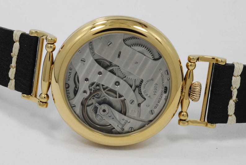 May include: A gold pocket watch with a black leather strap and a white stitching detail. The watch face is open, revealing the intricate inner workings of the watch. The watch is engraved with the text "E. Howard Watch Co. Boston, Mass. 02864".