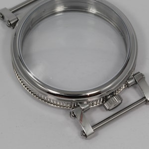 May include: A close-up of a shiny, silver-colored watch case with a clear glass face. The case has a circular design with a detailed, textured edge and hinged metal bars for attachment. The watch case is designed to hold a watch face.