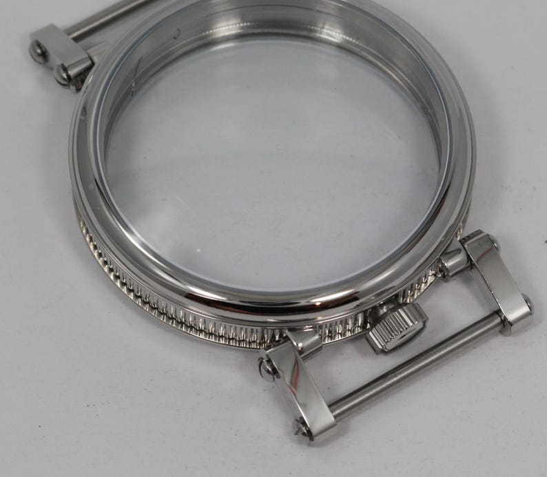 May include: A close-up of a shiny, silver-colored watch case with a clear glass face. The case has a circular design with a detailed, textured edge and hinged metal bars for attachment. The watch case is designed to hold a watch face.