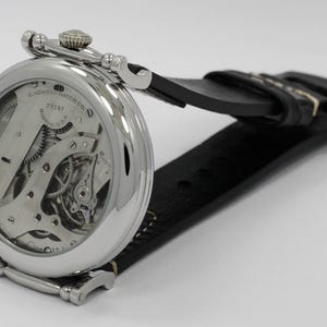 K&ouml;nnte beinhalten: Eine silberfarbene Armbanduhr mit poliertem, rundem Geh&auml;use und schwarzem Lederarmband. Die Uhr verf&uuml;gt &uuml;ber ein offenes mechanisches Uhrwerk, das durch einen transparenten Geh&auml;useboden sichtbar ist, mit dem Aufdruck E. HOWARD WATCH CO. BOSTON U.S.A.