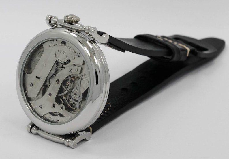 K&ouml;nnte beinhalten: Eine silberfarbene Armbanduhr mit poliertem, rundem Geh&auml;use und schwarzem Lederarmband. Die Uhr verf&uuml;gt &uuml;ber ein offenes mechanisches Uhrwerk, das durch einen transparenten Geh&auml;useboden sichtbar ist, mit dem Aufdruck E. HOWARD WATCH CO. BOSTON U.S.A.