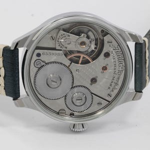 May include: A silver-toned wristwatch with a visible mechanical movement and a leather strap. The watch face displays intricate gears, springs, and the text "A.W.Co. Riverside, Waltham, Mass." The watch has a serial number 6559989.
